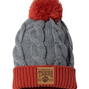May include: Gray cable knit beanie with a red pom pom and a brown leather patch that says "ARMADA TIGERS" with a paw print.