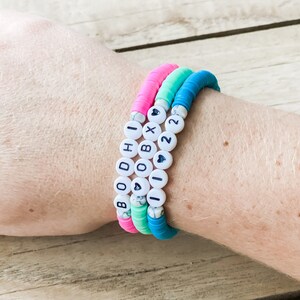 Puede incluir: Tres pulseras coloridas apiladas en una muñeca. Las pulseras son rosa, turquesa y azul. Una pulsera tiene cuentas de letras blancas que deletrean "OBX 22". Las pulseras están hechas de pequeñas cuentas redondas y planas.