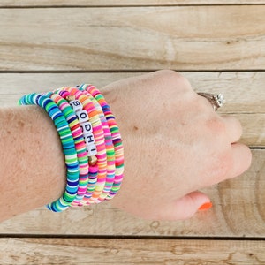 May include: A stack of colorful beaded bracelets on a wrist. The bracelets feature a variety of bright colors, including blue, green, pink, orange, and yellow. One bracelet spells out the word "BODHI" with white letter beads. The background is a wooden surface.