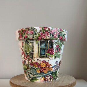 May include: A tapered cylindrical plant pot featuring a decoupage design of a rustic stone cottage exterior. The surface displays vibrant pink climbing roses, a blue window frame, and a green wheelbarrow overflowing with colorful autumn flowers.