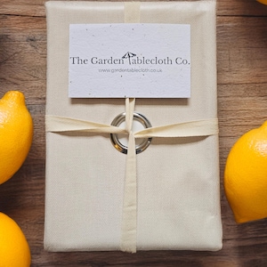 Op de afbeelding: Een rechthoekig, beige tafelkleed, verpakt en vastgebonden met een crèmekleurig lint. Een witte kaart met de tekst "The Garden Tablecloth Co." is bevestigd. Achtergrond: citroenen en een houten oppervlak.