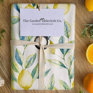 May include: A wrapped tablecloth featuring a lemon and leaf design. The white fabric is adorned with yellow lemons and green and blue leaves. A tag with the text "The Garden Tablecloth Co." is attached with a cream ribbon. The tablecloth is surrounded by lemons and oranges.