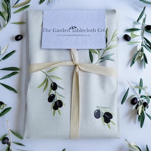 Könnte beinhalten: Ein cremefarbenes Tischtuch, gebunden mit einem beigen Band, zeigt Olivenzweig-Illustrationen und den Text "The Garden Tablecloth Co." Olivenzweige und Oliven sind um das verpackte Tischtuch verstreut.
