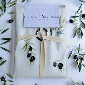 Könnte beinhalten: Eine gefaltete, cremefarbene Tischdecke mit einem aufgedruckten Olivenzweig-Design, gebunden mit einem cremefarbenen Band und einem silbernen Ring. Das Etikett lautet "The Garden Tablecloth Co." Olivenzweige und Oliven sind um die Tischdecke verstreut.