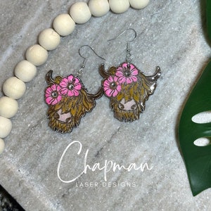 May include: A pair of silver earrings featuring a Highland cow head with pink flowers. The cow has brown fur and a pink nose.
