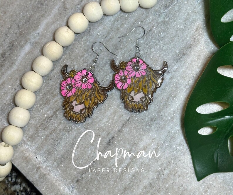 May include: A pair of silver earrings featuring a Highland cow head with pink flowers. The cow has brown fur and a pink nose.