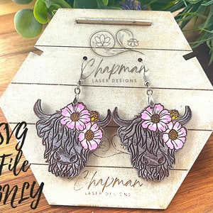 May include: A pair of wooden earrings featuring a Highland Cow design with pink flowers. The earrings are on a wooden display stand with the text "Chapman Laser Designs".