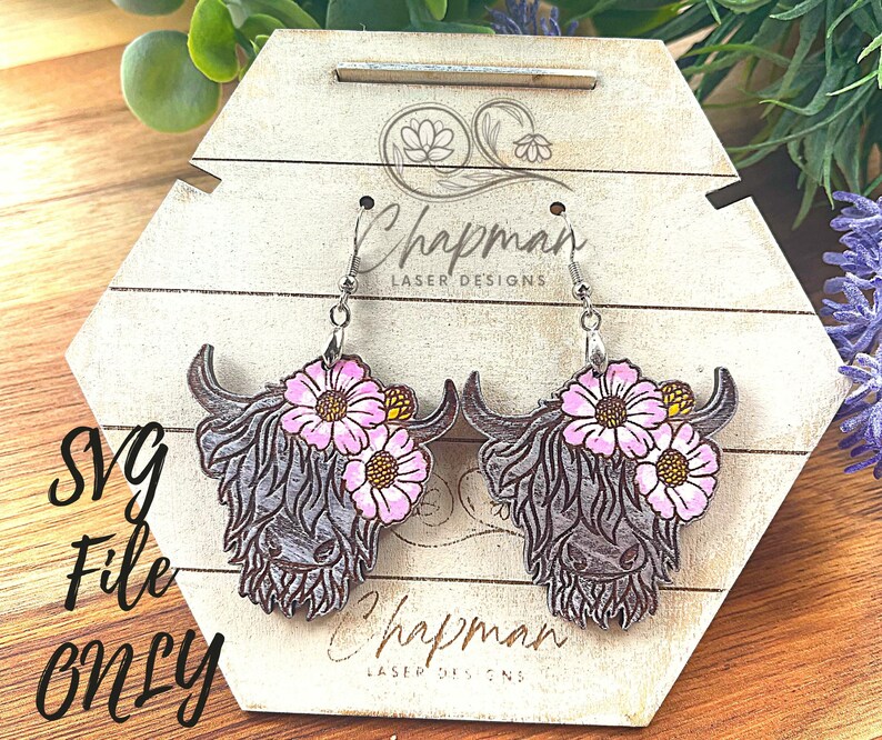 May include: A pair of wooden earrings featuring a Highland Cow design with pink flowers. The earrings are on a wooden display stand with the text "Chapman Laser Designs".