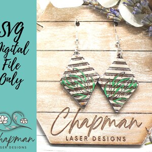 Op de afbeelding: Een paar diamantvormige oorbellen met een witte en bruine gestreepte achtergrond en groene bladeren. De oorbellen hangen aan zilveren haken op een houten achtergrond met de tekst "Chapman Laser Designs".