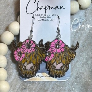 May include: Sterling silver earrings featuring a Highland cow with pink flowers and a sparkly rhinestone accent. The earrings are handmade in Idaho and are labelled "Chapman Laser Designs".