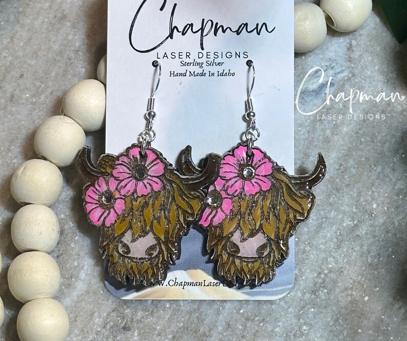 May include: Sterling silver earrings featuring a Highland cow with pink flowers and a sparkly rhinestone accent. The earrings are handmade in Idaho and are labelled "Chapman Laser Designs".