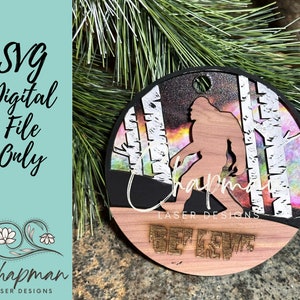 Peut inclure: Une décoration ronde en bois avec un fond noir et une silhouette de Bigfoot marchant dans une forêt de bouleaux. La décoration a une finition scintillante et irisée et les mots "Believe" et "Chapman Laser Designs" sont imprimés sur le devant.