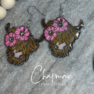 May include: A pair of earrings featuring a Highland cow with pink flowers. The earrings are made of wood and have a shiny, glossy finish.