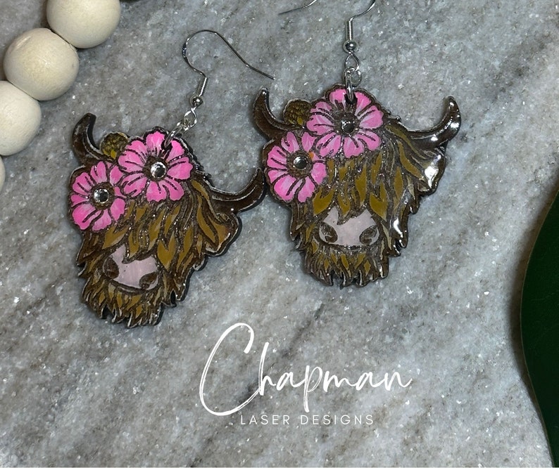 May include: A pair of earrings featuring a Highland cow with pink flowers. The earrings are made of wood and have a shiny, glossy finish.