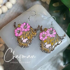 May include: A pair of silver earrings featuring a Highland cow with pink flowers. The earrings are made of metal and have a shiny finish.