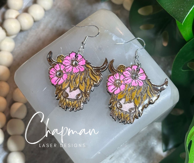 May include: A pair of silver earrings featuring a Highland cow with pink flowers. The earrings are made of metal and have a shiny finish.