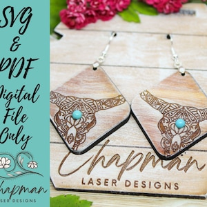 May include: A pair of wooden earrings with a laser-cut design of a cow skull with turquoise accents. The earrings are hanging from silver hooks and are displayed on a wooden surface with the text "Chapman Laser Designs".