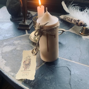 May include: A beige candle with a gold rune carved into it. The candle is tied with twine and has a label that reads "Honoring Loki".