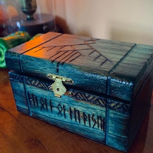 May include: A wooden box with a teal finish and intricate carvings. The box features a brass clasp and is adorned with runes. The lid has a carved design, and the box is set on a wooden surface. The overall aesthetic is rustic and detailed.