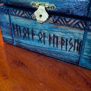 May include: A wooden chest with a dark blue finish and brass hardware. The chest features carved runic inscriptions and decorative patterns. The lid is closed with a brass clasp. The chest is on a wooden surface.