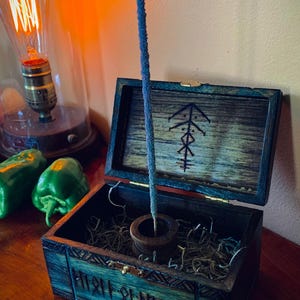 May include: A blue incense stick stands in a wooden holder inside an open, teal-colored wooden box. The box has a carved design and a symbol on the lid. The box sits on a wooden surface, with a vintage light bulb and green peppers in the background.