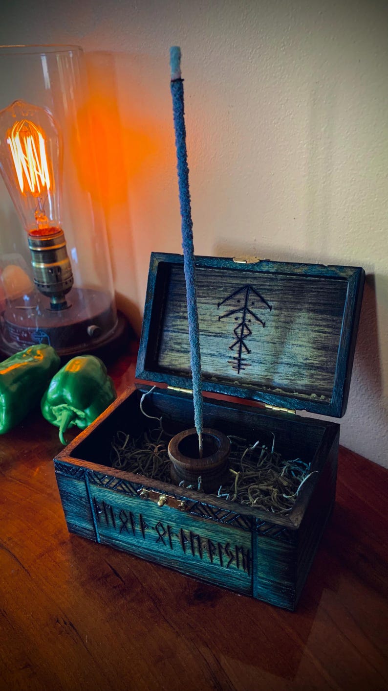 May include: A blue incense stick stands in a wooden holder inside an open, teal-colored wooden box. The box has a carved design and a symbol on the lid. The box sits on a wooden surface, with a vintage light bulb and green peppers in the background.