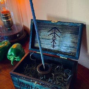 May include: An open wooden box with a blue-green finish, containing dried moss and a small wooden incense holder. A blue incense stick stands upright. The box lid is decorated with a black symbol. A vintage light bulb and green peppers are in the background.