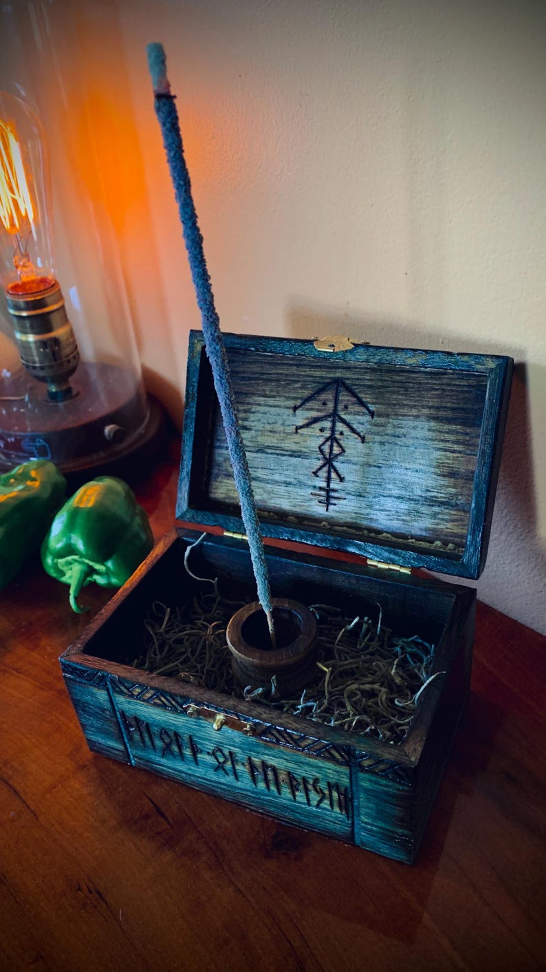 May include: An open wooden box with a blue-green finish, containing dried moss and a small wooden incense holder. A blue incense stick stands upright. The box lid is decorated with a black symbol. A vintage light bulb and green peppers are in the background.
