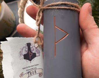 May include: Gray candle with a copper-colored rune symbol on the front. The candle is tied with twine and has a tag that says "Where the Moon Meets the Sun, Honoring Thor".
