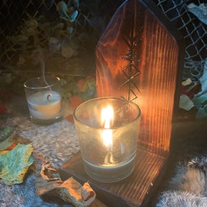 K&ouml;nnte beinhalten: Ein Kerzenhalter aus Holz mit einem geschnitzten Symbol, mit einer brennenden Kerze in einem Glasgef&auml;&szlig;. Der Halter steht auf einer flauschigen Oberfl&auml;che mit Bl&auml;ttern, die um ihn herum verstreut sind.