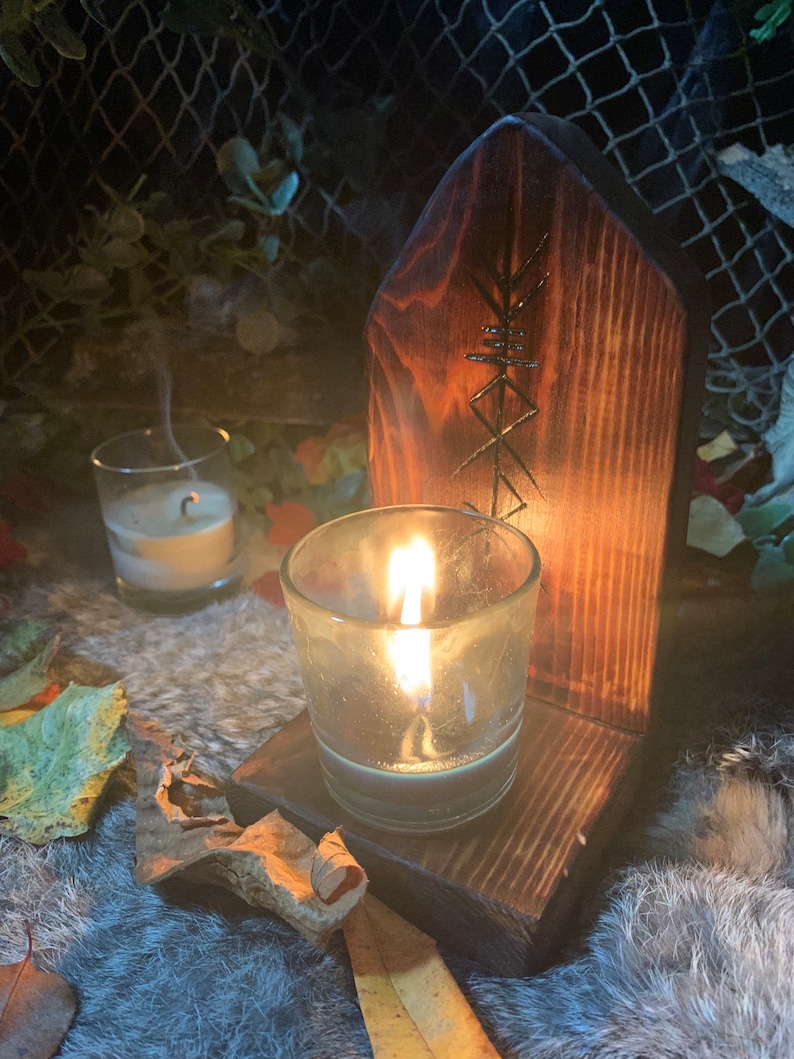 K&ouml;nnte beinhalten: Ein Kerzenhalter aus Holz mit einem geschnitzten Symbol, mit einer brennenden Kerze in einem Glasgef&auml;&szlig;. Der Halter steht auf einer flauschigen Oberfl&auml;che mit Bl&auml;ttern, die um ihn herum verstreut sind.