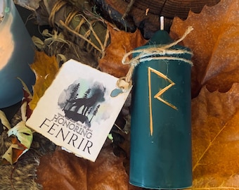 May include: A teal green candle with a gold rune symbol on the front. The candle is surrounded by fall leaves and a tag that reads "Honoring Fenrir".