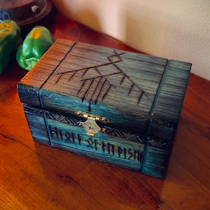 May include: A wooden box with a dark teal and brown finish. The box features carved designs on the lid and front, with a brass clasp. The box is rectangular and appears to be handcrafted, with a rustic aesthetic.