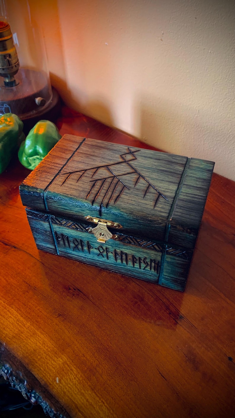 May include: A wooden box with a dark teal and brown finish. The box features carved designs on the lid and front, with a brass clasp. The box is rectangular and appears to be handcrafted, with a rustic aesthetic.