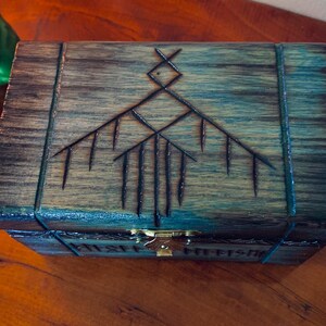 May include: Wooden box with a dark finish, featuring an etched geometric design on the lid. The box has a brass clasp and is placed on a wooden surface. The design is a series of lines and shapes.