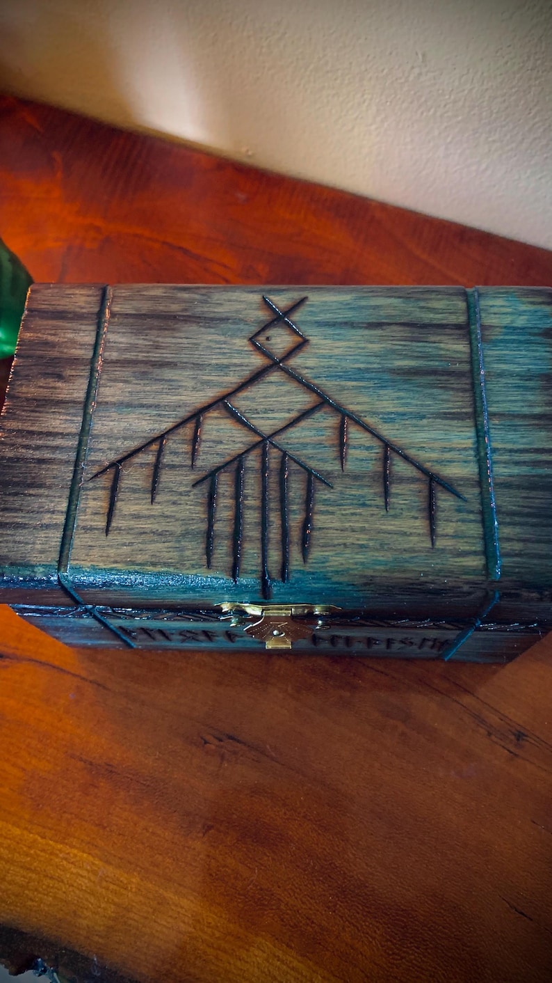 May include: Wooden box with a dark finish, featuring an etched geometric design on the lid. The box has a brass clasp and is placed on a wooden surface. The design is a series of lines and shapes.