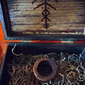 May include: Open wooden box with a carved design on the lid. Inside, a wooden cylinder sits atop a bed of dried plant material. The box is stained a dark brown color.