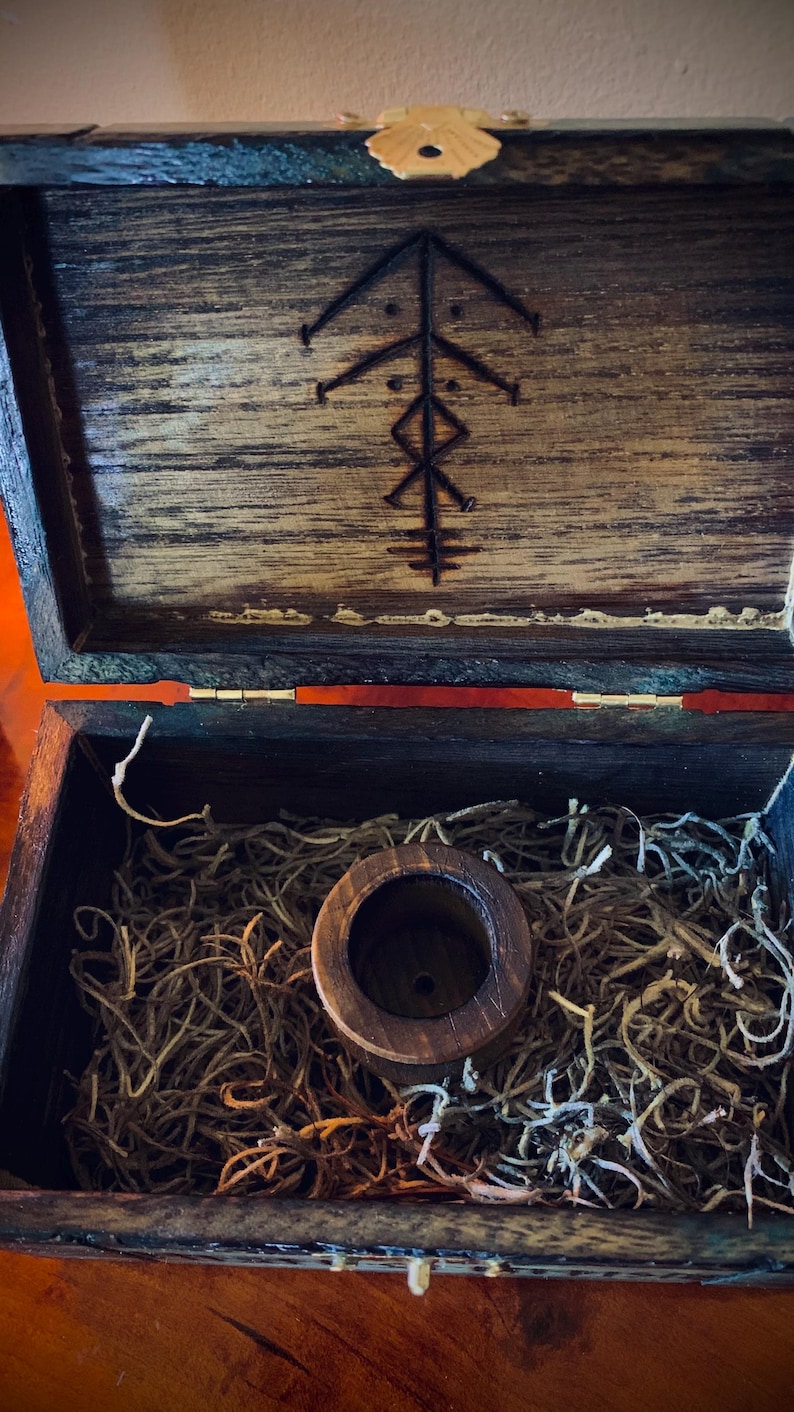 May include: Open wooden box with a carved design on the lid. Inside, a wooden cylinder sits atop a bed of dried plant material. The box is stained a dark brown color.