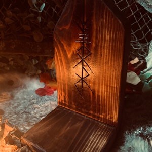 K&ouml;nnte beinhalten: Ein Holzaltar mit einem geschnitzten Symbol, m&ouml;glicherweise einer Rune, auf der Vorderseite. Der Altar ist dunkelbraun und hat eine raue, rustikale Textur. Er steht auf einer wei&szlig;en, flauschigen Oberfl&auml;che.