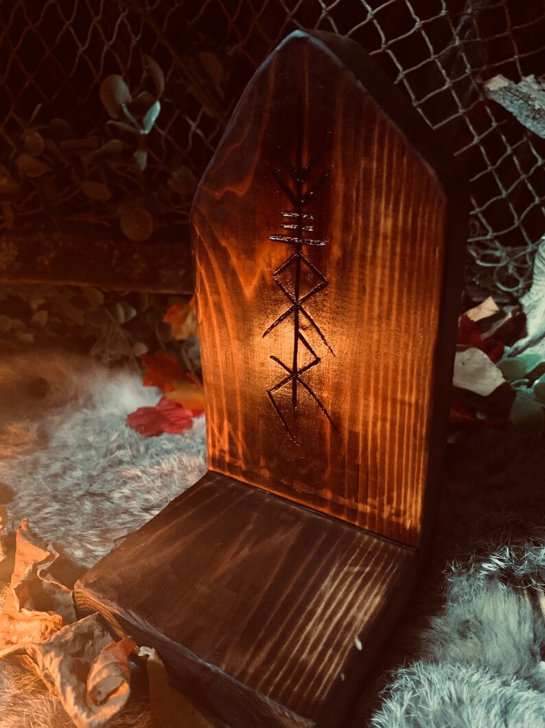 K&ouml;nnte beinhalten: Ein Holzaltar mit einem geschnitzten Symbol, m&ouml;glicherweise einer Rune, auf der Vorderseite. Der Altar ist dunkelbraun und hat eine raue, rustikale Textur. Er steht auf einer wei&szlig;en, flauschigen Oberfl&auml;che.