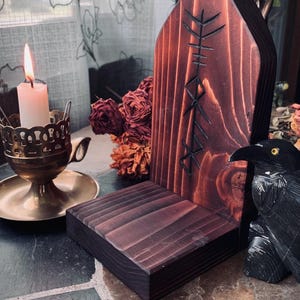 K&ouml;nnte beinhalten: Ein dunkelbrauner Holzaltar mit einem geschnitzten Symbol auf der Vorderseite. Der Altar hat eine kleine Ablage und steht auf einer gefliesten Oberfl&auml;che. Eine schwarze Rabenfigur befindet sich rechts vom Altar.