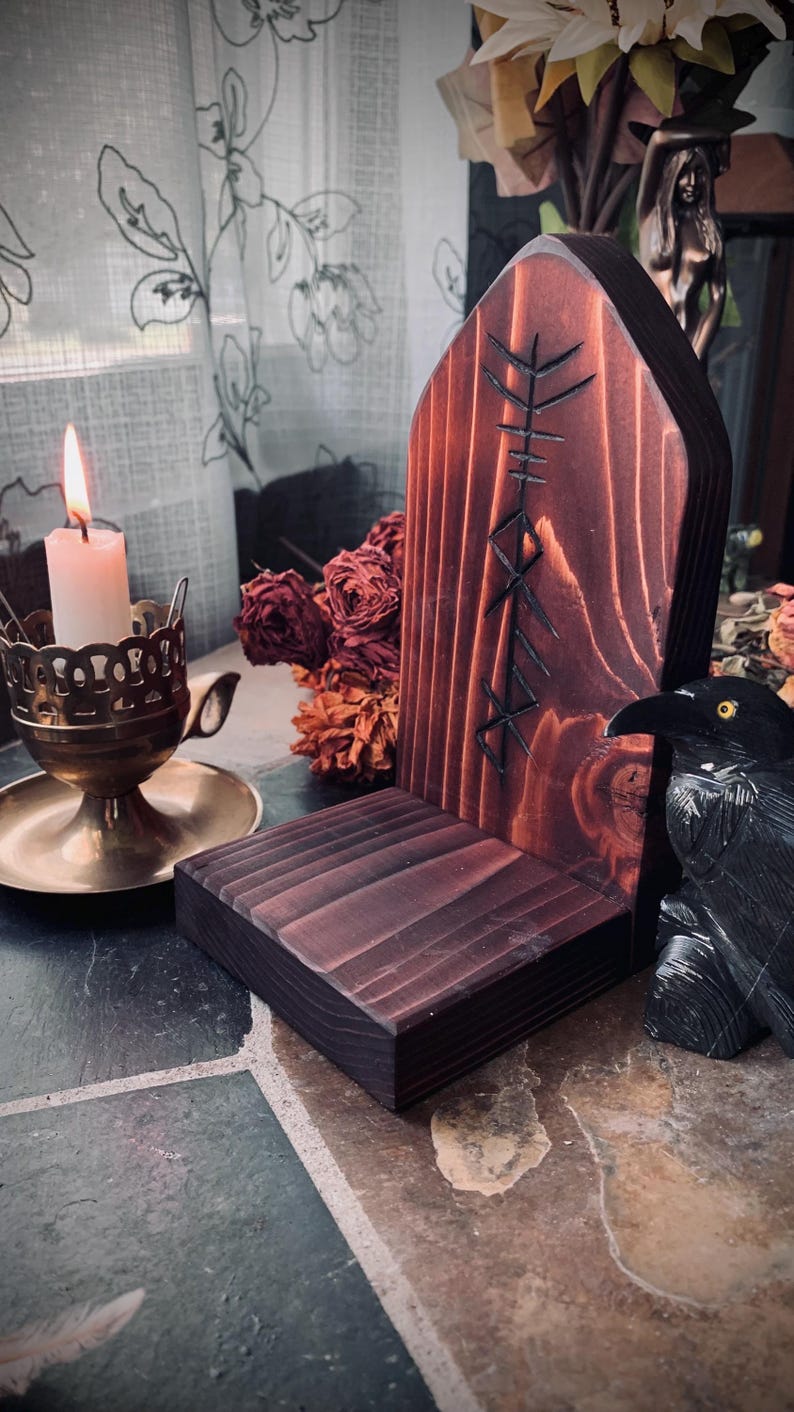 K&ouml;nnte beinhalten: Ein dunkelbrauner Holzaltar mit einem geschnitzten Symbol auf der Vorderseite. Der Altar hat eine kleine Ablage und steht auf einer gefliesten Oberfl&auml;che. Eine schwarze Rabenfigur befindet sich rechts vom Altar.