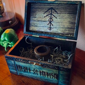 May include: Open wooden box with a dark teal exterior and a lighter wood interior. The lid is open, revealing a carved symbol. Inside, a small wooden cylinder sits on a bed of dried grass. The box is on a wooden surface.