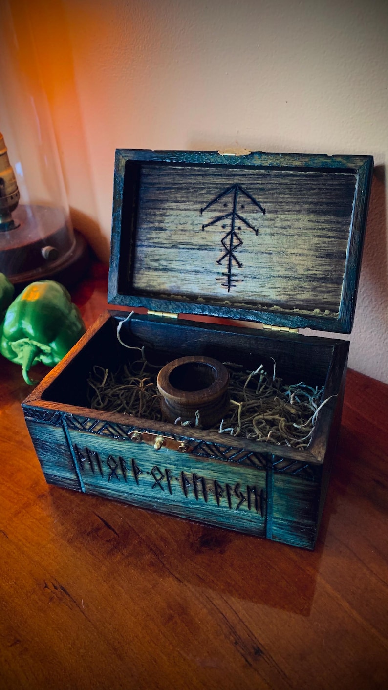 May include: Open wooden box with a dark teal exterior and a lighter wood interior. The lid is open, revealing a carved symbol. Inside, a small wooden cylinder sits on a bed of dried grass. The box is on a wooden surface.