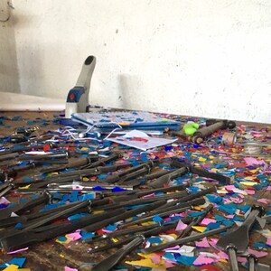 May include: A messy workbench with a variety of tools, including a hole punch, a hammer, and many small, sharp tools. The workbench is covered in colorful paper scraps.