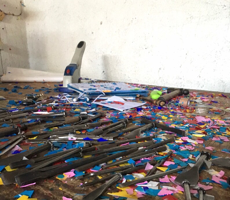 May include: A messy workbench with a variety of tools, including a hole punch, a hammer, and many small, sharp tools. The workbench is covered in colorful paper scraps.