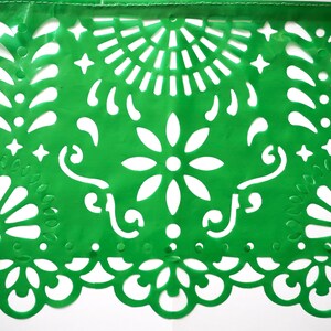May include: Green paper banner with a traditional Mexican design featuring cut-out flowers, stars, and swirls. The banner is made of thin paper and has a delicate, intricate design.