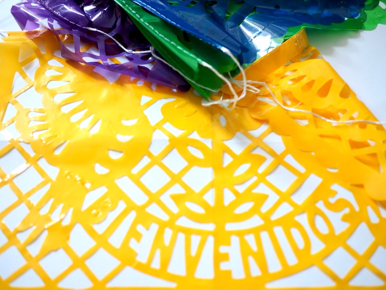 May include: A yellow paper banner with a cut-out design featuring the words "Bienvenidos" in Spanish. The banner is part of a string of colorful paper banners.