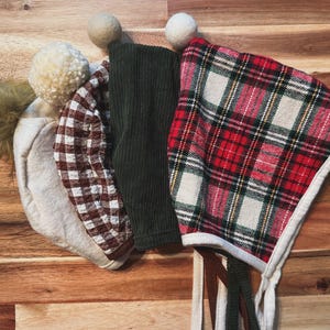 May include: A collection of baby bonnets in various styles and colors. One bonnet features a red and green plaid pattern, another has a brown and white checkered design, and a third is a solid dark green corduroy. Each bonnet has a unique trim.
