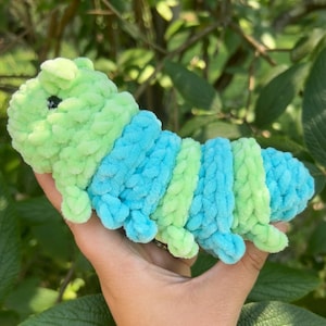 Puede incluir: Un juguete de oruga de peluche hecho a mano en tonos verdes y azules. La oruga tiene un cuerpo segmentado con bandas alternas de verde claro y azul. Tiene patas pequeñas y rechonchas y un pequeño ojo negro. El juguete está hecho de un material suave y texturizado.